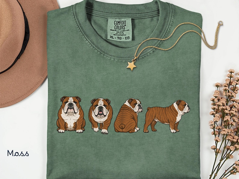 May include: A moss green t-shirt with a series of four cartoon bulldogs in different poses. The shirt has a crew neck and short sleeves. A gold-coloured necklace with a star pendant hangs on the shirt. The text "COMFORT COLORS" is visible on the tag.