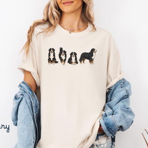 Op de afbeelding: Ivoor kleurig T-shirt met een afbeelding van Berner Sennenhonden in verschillende poses. De honden zijn afgebeeld in zwart, wit en bruin. Het woord "Ivory" is in handschriftstijl links onderaan het shirt gedrukt.
