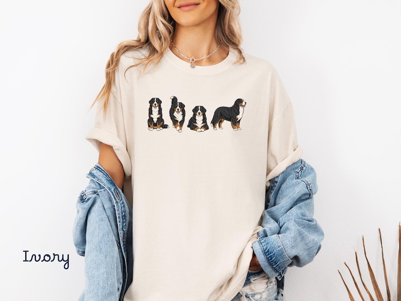 Op de afbeelding: Ivoor kleurig T-shirt met een afbeelding van Berner Sennenhonden in verschillende poses. De honden zijn afgebeeld in zwart, wit en bruin. Het woord "Ivory" is in handschriftstijl links onderaan het shirt gedrukt.