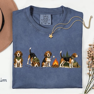 May include: A blue t-shirt featuring a graphic of four beagle dogs in various poses, including sitting, sniffing the ground, roasting a marshmallow over a campfire, and standing near a tent and pine trees. The shirt has a Comfort Colors label.