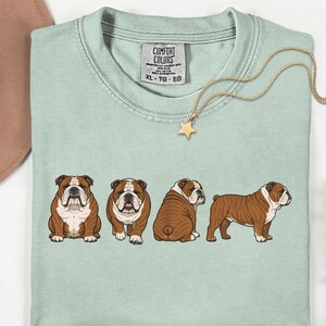May include: A pale green t-shirt featuring a graphic of four brown and white bulldogs in various poses. A gold necklace with a star pendant and a brown hat are also visible. The shirt has the text "Comfort Colors" on the tag.