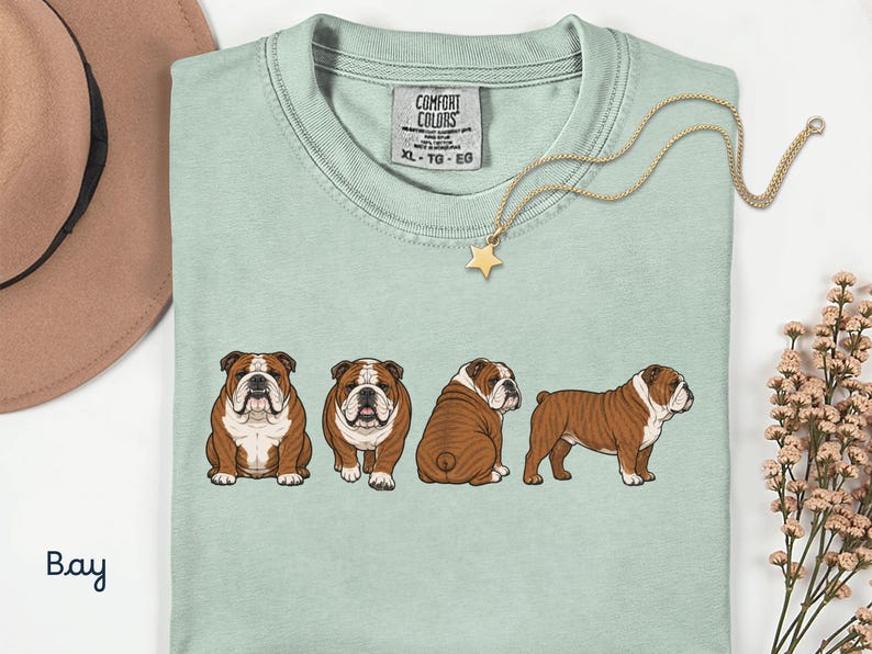 May include: A pale green t-shirt featuring a graphic of four brown and white bulldogs in various poses. A gold necklace with a star pendant and a brown hat are also visible. The shirt has the text "Comfort Colors" on the tag.