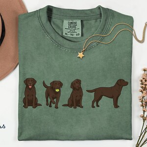 K&ouml;nnte beinhalten: Ein moosgr&uuml;nes T-Shirt mit einer Grafik von vier braunen Labrador-Retrievern in verschiedenen Posen, darunter einer mit einem Tennisball. Das Shirt hat ein Comfort Colors-Etikett am Kragen, Gr&ouml;&szlig;e XL, und wird mit einem beigen Hut und einer Sternkette gezeigt.