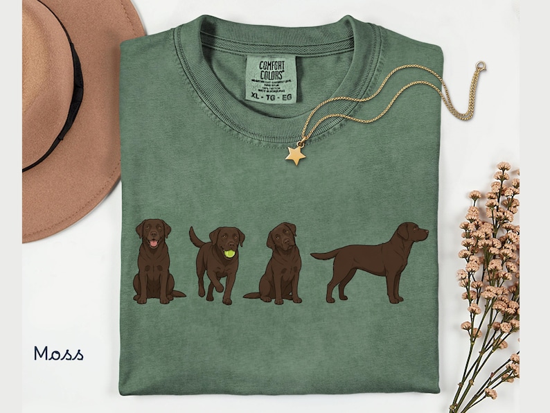 K&ouml;nnte beinhalten: Ein moosgr&uuml;nes T-Shirt mit einer Grafik von vier braunen Labrador-Retrievern in verschiedenen Posen, darunter einer mit einem Tennisball. Das Shirt hat ein Comfort Colors-Etikett am Kragen, Gr&ouml;&szlig;e XL, und wird mit einem beigen Hut und einer Sternkette gezeigt.