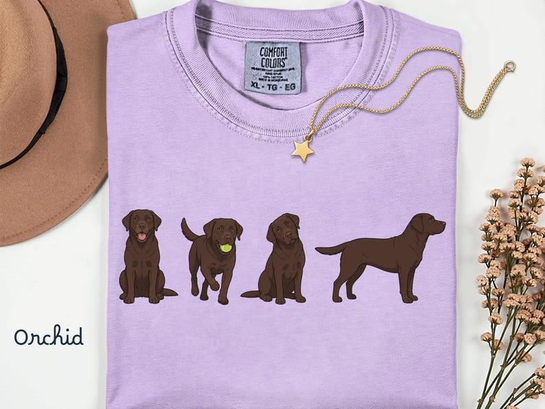 K&ouml;nnte beinhalten: Ein hellviolettes T-Shirt mit einer Grafik von vier schokoladenbraunen Labradoren in verschiedenen Posen, darunter einer mit einem Tennisball. Das Shirt hat ein Comfort Colors-Etikett am Kragen mit der Gr&ouml;&szlig;e XL-TG-EG und dem Wort Orchid darunter.