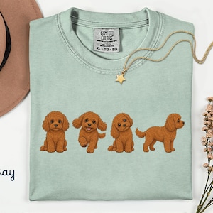 May include: A light green crew neck t-shirt featuring a graphic print of four brown curly-haired dogs in different poses. The shirt has a Comfort Colors brand label at the inner neckline with size XL, TG, EG printed. The word Bay is printed in blue.