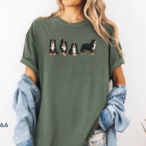 Op de afbeelding: Mosgroen t-shirt met een geborduurd ontwerp van vier Berner Sennenhonden in verschillende poses. De honden zijn zwart, wit en bruin. Het shirt heeft korte mouwen en een relaxte pasvorm. Het woord "Moss" staat linksonder gedrukt.