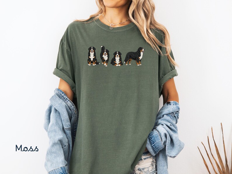Op de afbeelding: Mosgroen t-shirt met een geborduurd ontwerp van vier Berner Sennenhonden in verschillende poses. De honden zijn zwart, wit en bruin. Het shirt heeft korte mouwen en een relaxte pasvorm. Het woord "Moss" staat linksonder gedrukt.