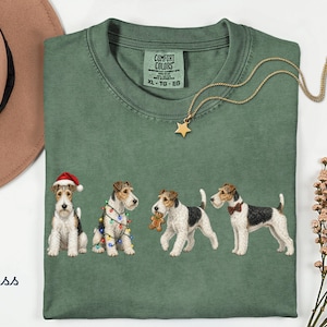 Könnte beinhalten: Ein moosgrünes T-Shirt mit einer Grafik von vier Foxterriern. Ein Hund trägt eine Weihnachtsmannmütze, einer ist mit bunten Lichterketten umwickelt, einer hält einen Lebkuchenmann und einer trägt eine Fliege. Das Shirt hat ein Comfort Colors Etikett.