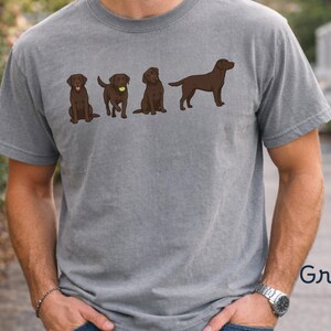 K&ouml;nnte beinhalten: Ein graues, kurz&auml;rmeliges T-Shirt mit einem Grafikdruck von vier braunen Labrador-Retriever-Hunden in verschiedenen Posen, darunter einer mit einem gelben Tennisball. Das T-Shirt hat einen Rundhalsausschnitt und das Wort Grey ist aufgedruckt.