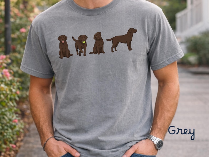 K&ouml;nnte beinhalten: Ein graues, kurz&auml;rmeliges T-Shirt mit einem Grafikdruck von vier braunen Labrador-Retriever-Hunden in verschiedenen Posen, darunter einer mit einem gelben Tennisball. Das T-Shirt hat einen Rundhalsausschnitt und das Wort Grey ist aufgedruckt.