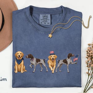 May include: A denim blue t-shirt featuring a graphic of four dogs. One golden retriever wears a blue star-patterned bandana, a German shorthaired pointer wears a rosette, another golden retriever wears a red, white, and blue top hat, and a second pointer holds a small American flag.