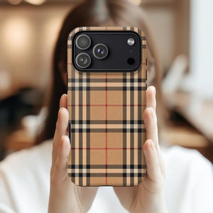 May include: A phone case with a tan, black, and red plaid pattern. The case has a black camera module with three lenses and a flash. The phone case is held in front of a person.