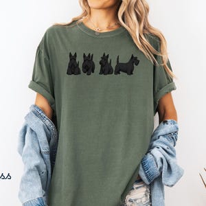 May include: Moss green t-shirt featuring a line of four black Scottish Terrier dog illustrations. The shirt has a relaxed fit with rolled-up sleeves. The word "Moss" is printed in white on the lower left side of the image.