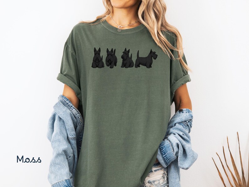 May include: Moss green t-shirt featuring a line of four black Scottish Terrier dog illustrations. The shirt has a relaxed fit with rolled-up sleeves. The word "Moss" is printed in white on the lower left side of the image.
