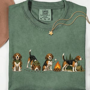 Könnte beinhalten: Ein moosgrünes T-Shirt mit einer Grafik von vier Beagle-Hunden beim Camping. Die Hunde sind sitzend, mit einem Hut am Boden schnüffelnd, beim Rösten eines Marshmallows über einem Lagerfeuer und stehend neben einem Zelt und Tannen abgebildet.