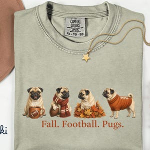 Peut inclure: Un t-shirt à col rond kaki avec un motif représentant quatre carlins dans des décors d'automne, dont un avec une tasse, un avec une écharpe et un ballon, un dans des feuilles mortes et un avec un pull. Le texte indique Fall. Football. Pugs.