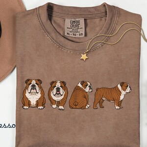 May include: Espresso-coloured t-shirt with a crew neck, featuring a graphic of four English Bulldogs in different poses. A gold necklace with a star pendant and a brown hat are also visible.