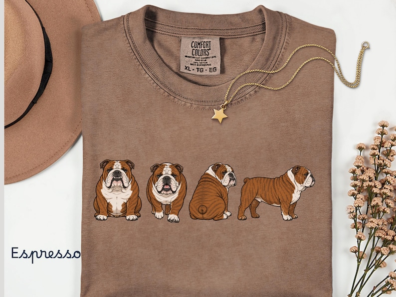 May include: Espresso-coloured t-shirt with a crew neck, featuring a graphic of four English Bulldogs in different poses. A gold necklace with a star pendant and a brown hat are also visible.