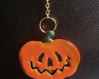 May include: A Halloween-themed keychain featuring an orange pumpkin charm with a carved face. The pumpkin is attached to a gold-colored chain and keyring. The background is a textured black surface.