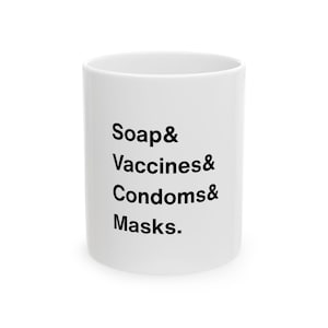 May include: A white ceramic mug with black text that reads "Soap & Vaccines & Condoms & Masks." The mug is cylindrical and has a simple, clean design. The text is vertically aligned on the side of the mug.