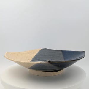 May include: A shallow ceramic bowl featuring a tri-color design with a textured beige section, a matte dark gray middle, and a smooth glossy blue edge. The bowl has a wide, slightly curved rim and sits on a raised, circular base with a light tan finish.