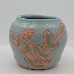 May include: A small, round, light blue ceramic vase with a floral design. The design features pink and green leaves with orange berries. The vase has a smooth, glossy finish and a wide opening. A decorative piece for the home.