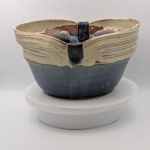 May include: A ceramic bowl with a wide, undulating rim featuring a cream-colored upper section and a deep blue lower section. The bowl has a dark, vertical handle attachment at the center of the rim, showcasing a two-tone glaze finish.