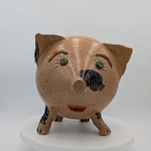 May include: A ceramic pig-shaped planter with a tan speckled finish. The pig has large ears, green eyes, and a black patch on its snout. The pig has a red smiling mouth and black and tan legs. The planter is on a white pedestal.