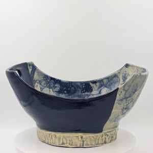 May include: A ceramic bowl with a curved, asymmetrical rim and two integrated handles. The exterior features a smooth, deep navy blue glaze on one side, contrasting with a textured, light beige base and a blue bubble-patterned glaze finish.