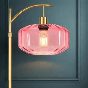 K&ouml;nnte beinhalten: Eine Stehlampe mit einem St&auml;nder aus geb&uuml;rstetem Goldmetall, einem geschwungenen Arm und einem breiten, durchscheinenden rosa Glasschirm. Die Lampe enth&auml;lt eine einzelne Gl&uuml;hbirne mit sichtbarem Filament, die ein warmes Licht abgibt.