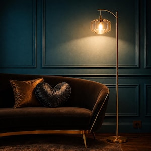 K&ouml;nnte beinhalten: Eine hohe, schlanke Stehlampe aus Messing mit einem geometrischen, facettierten Glasschirm, der ein warmes Licht ausstrahlt. Die Lampe hat einen geschwungenen Arm und einen runden Fu&szlig; und steht neben einem dunkelbraunen, geschwungenen Sofa.
