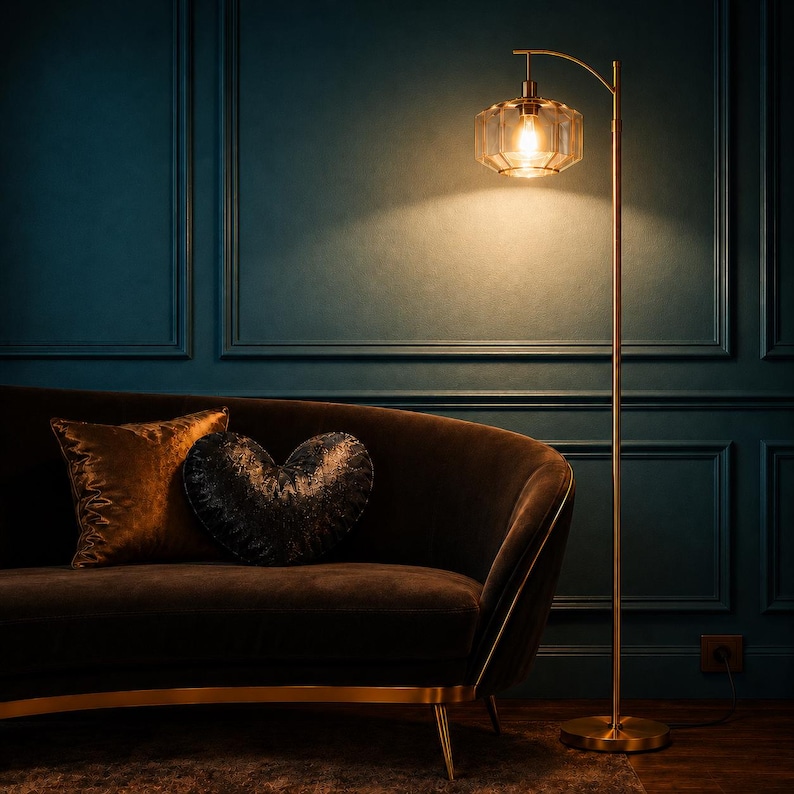 K&ouml;nnte beinhalten: Eine hohe, schlanke Stehlampe aus Messing mit einem geometrischen, facettierten Glasschirm, der ein warmes Licht ausstrahlt. Die Lampe hat einen geschwungenen Arm und einen runden Fu&szlig; und steht neben einem dunkelbraunen, geschwungenen Sofa.
