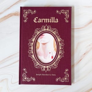 May include: A hardcover book titled Carmilla by Joseph Sheridan Le Fanu with a deep burgundy cover. The front features gold ornate filigree borders and a central oval frame containing an illustration of a neck with two small red blood drops.