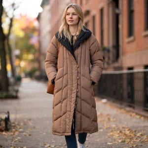 May include: A long, tan puffer coat featuring a black hood and collar lining, a button-front closure, and a quilted chevron pattern. The coat has long sleeves and reaches below the knees, providing full coverage with a structured, insulated design.