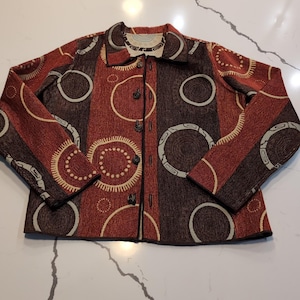 May include: A long-sleeved button-down jacket featuring a textured pattern of large circles in shades of rust orange, dark brown, and cream. The garment has a standard collar, a front button closure, and a repeating geometric circle motif throughout.