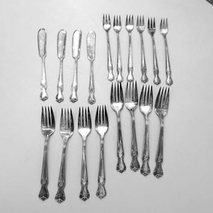 May include: A collection of sixteen silver-toned metal utensils consisting of four butter knives with rounded blades and twelve forks of varying sizes, all featuring ornate decorative detailing on the handles and polished metallic surfaces.