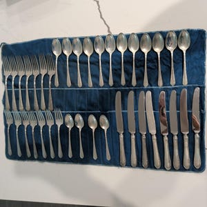 May include: A large set of silver-toned flatware arranged on a dark blue fabric roll. The collection includes twelve forks, twelve spoons of varying sizes, and eight knives, all featuring a simple, polished metal design with slender handles.