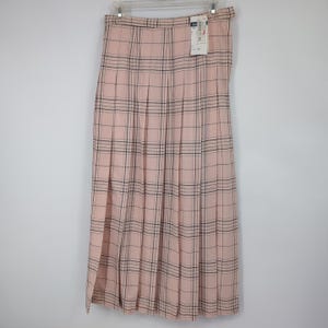 May include: A long, pleated skirt in a pink plaid pattern, hanging on a hanger. The skirt has a classic design with a high waist and a knee-length hem. The plaid pattern features thin black lines over the pink fabric. A tag is attached to the skirt.