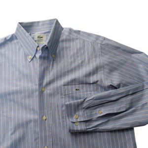 May include: A light blue and white striped long-sleeve button-down shirt featuring a button-down collar, a front chest pocket with a small green crocodile logo, and white buttons. The interior neck label displays the Lacoste brand name.