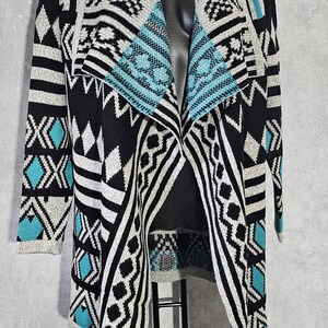 Pode incluir: Cardigã preto, branco e turquesa com frente aberta e padrão geométrico. O suéter de manga comprida apresenta gola xale e uma mistura de listras, losangos e ziguezagues. O cardigã é exibido em um manequim preto.