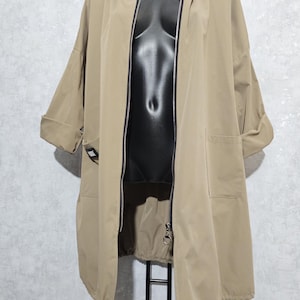 Könnte beinhalten: Beigefarbene Kapuzenjacke mit durchgehendem Reißverschluss, kurzen Ärmeln und zwei Vordertaschen. Die Jacke wird auf einer schwarzen Schaufensterpuppe präsentiert. Die Jacke hat einen Kordelzug am Saum und ist aus leichtem Material.