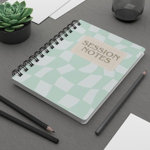 May include: A notebook with a spiral binding, featuring a light green and white chequered design. The cover has a beige label with the words "SESSION NOTES" in brown. Two black pencils and a small succulent plant are also visible.