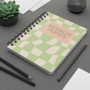 May include: A spiral-bound notebook with a green and cream checkered pattern. The cover features a peach-colored label with the words "SESSION NOTES" in brown. Two black pencils and a small succulent plant are also visible.