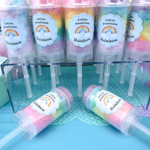 May include: A display of colorful cotton candy push pops. Each clear plastic container is filled with layers of rainbow-colored cotton candy and topped with a label that reads "Cotton Sweetness Rainbow."