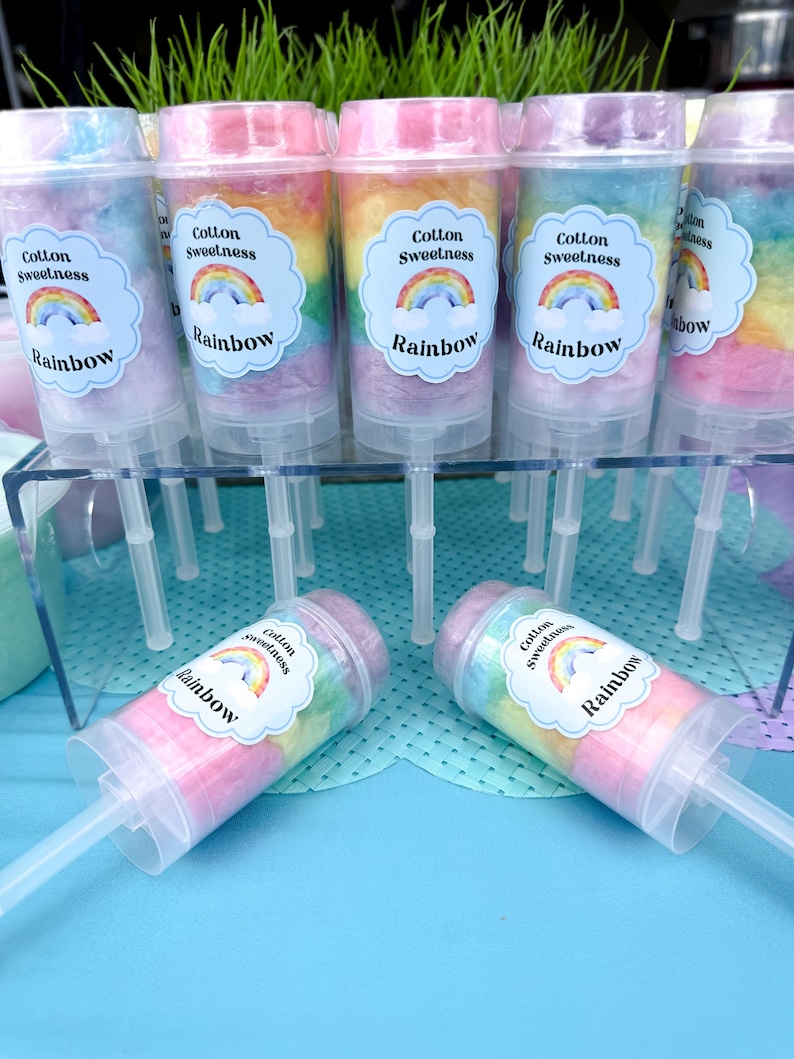 May include: A display of colorful cotton candy push pops. Each clear plastic container is filled with layers of rainbow-colored cotton candy and topped with a label that reads "Cotton Sweetness Rainbow."