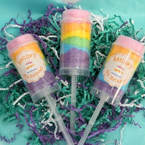 May include: Three push-pop containers filled with rainbow-colored cotton candy. Two containers have labels that read "Baylor's 5th Birthday" with a cake graphic. The containers are on a bed of teal and purple shredded paper.