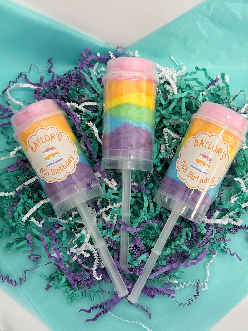 May include: Three push-pop containers filled with rainbow-colored cotton candy. Two containers have labels that read "Baylor's 5th Birthday" with a cake graphic. The containers are on a bed of teal and purple shredded paper.