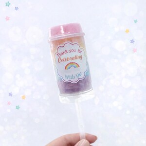 May include: A clear push pop container filled with rainbow-colored candy. The container has a pink lid and a label that reads "Thank you for Celebrating With Us!" A hand holds the clear plastic stick.