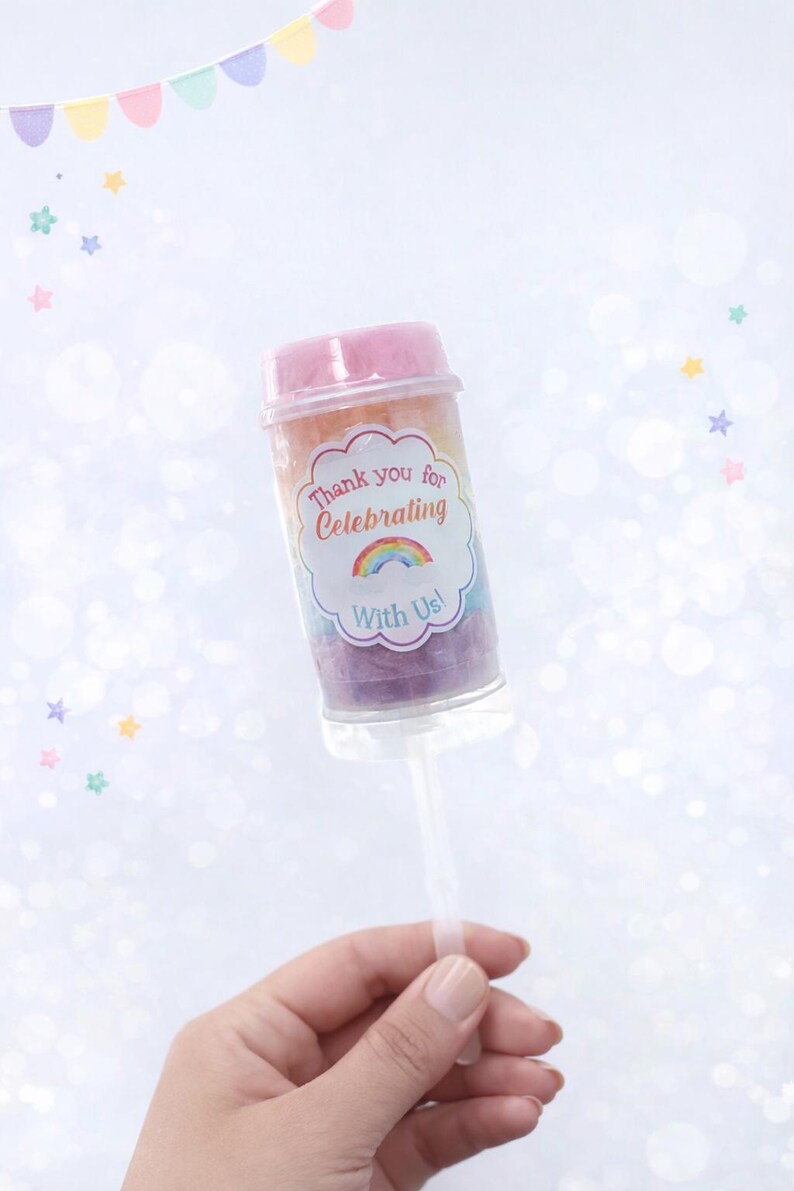 May include: A clear push pop container filled with rainbow-colored candy. The container has a pink lid and a label that reads "Thank you for Celebrating With Us!" A hand holds the clear plastic stick.